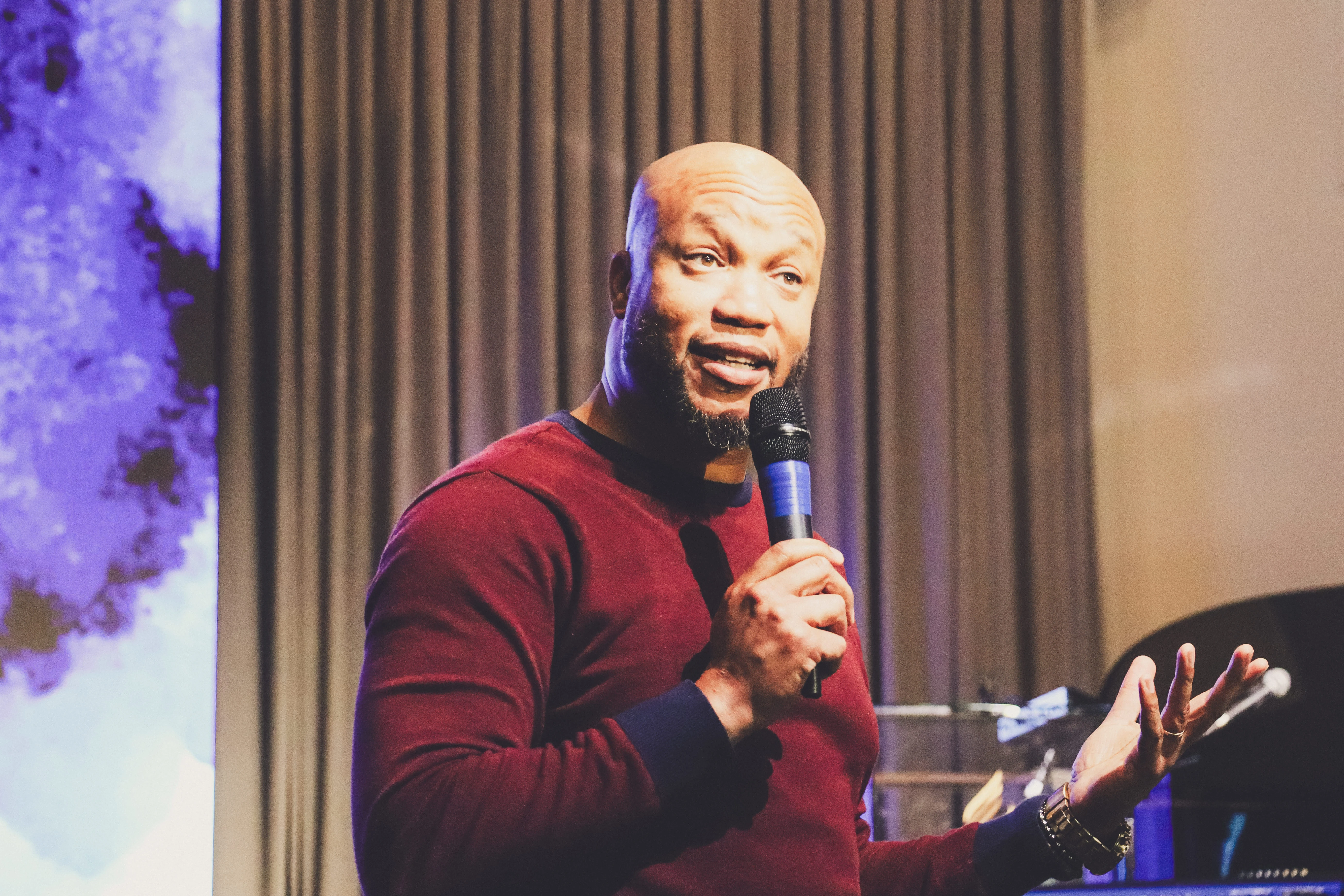 A man in a maroon sweater holds a microphone and speaks on stage, gesturing with one hand. Gray curtains and part of a grand piano are visible in the background.